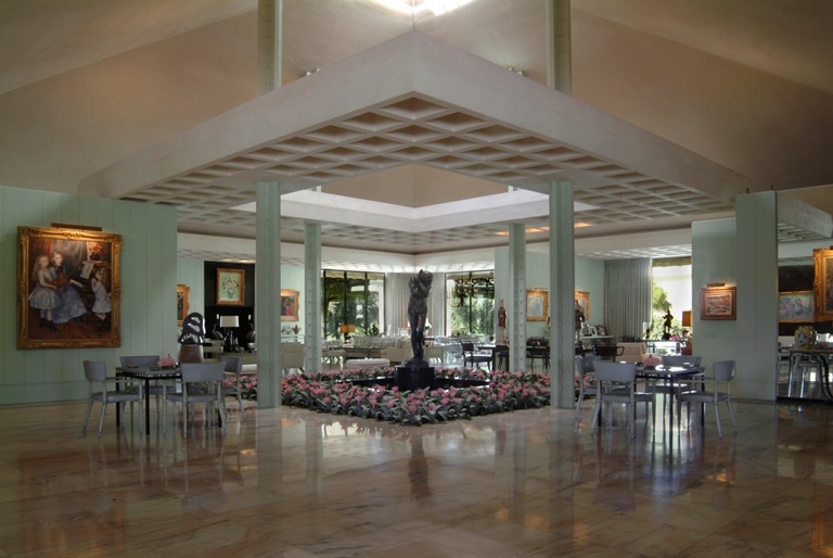 Elegant interior of a spacious room with marble floors, a central geometric ceiling pattern, and a series of square columns. The room features artwork on the walls, a large statue in the center surrounded by pink flowers, and multiple tables with chairs spread throughout.