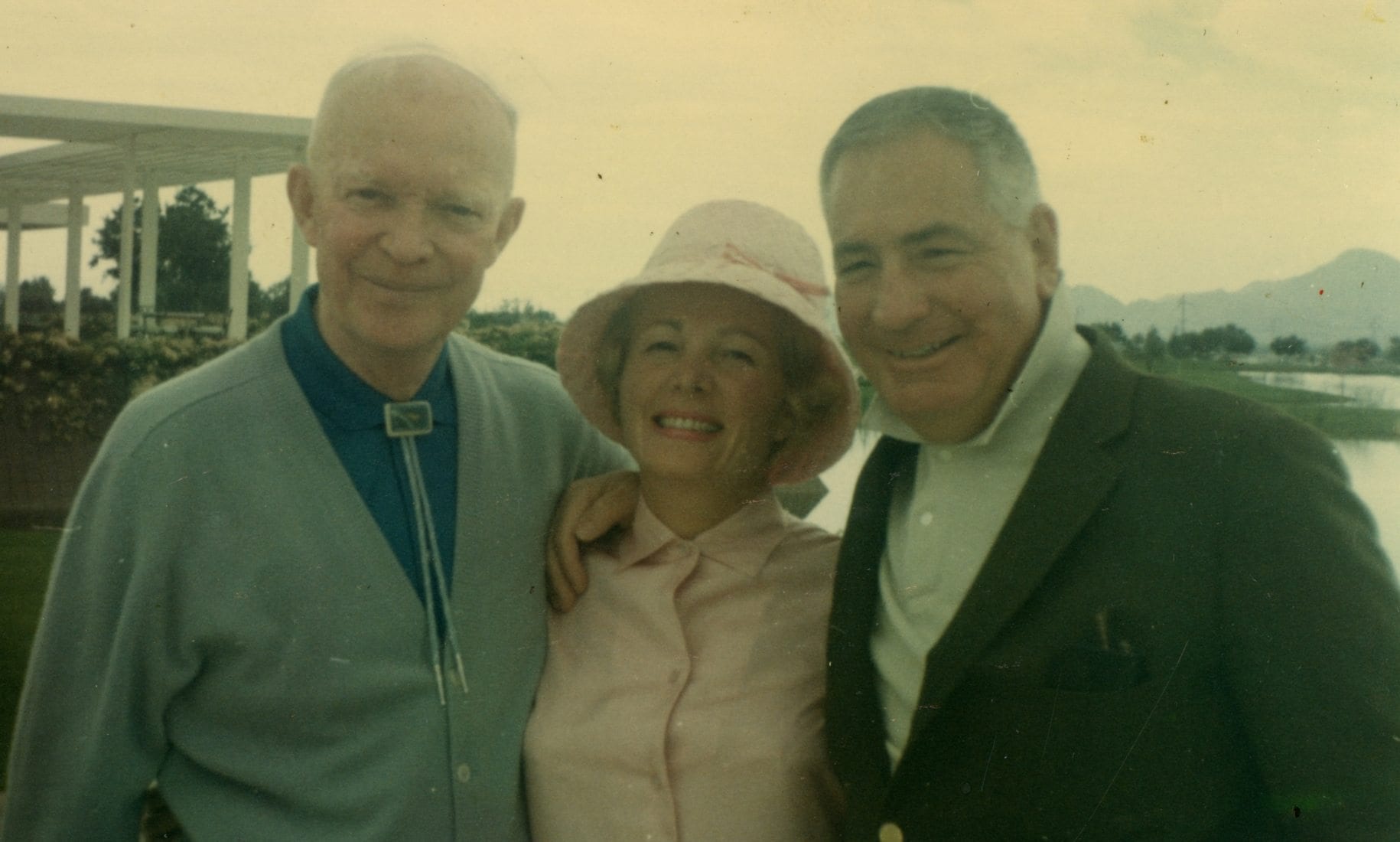 Three elderly individuals standing together outdoors, with a woman in the middle wearing a pink shirt and a wide-brimmed hat, flanked by two men in sweaters and jackets, with a gazebo and mountains in the background.