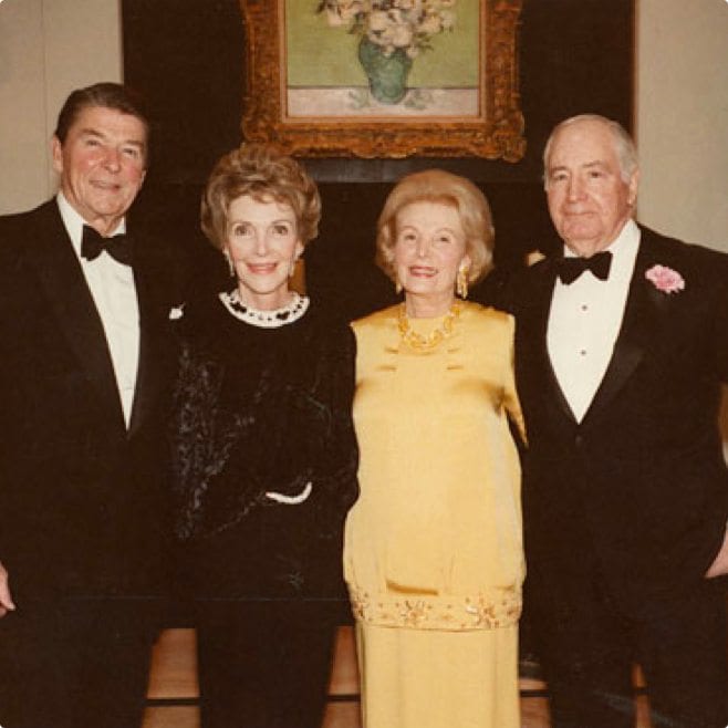 Four elderly individuals, two men wearing black tuxedos with bow ties and two women in elegant dresses, pose for a photo in a room with a framed painting in the background.