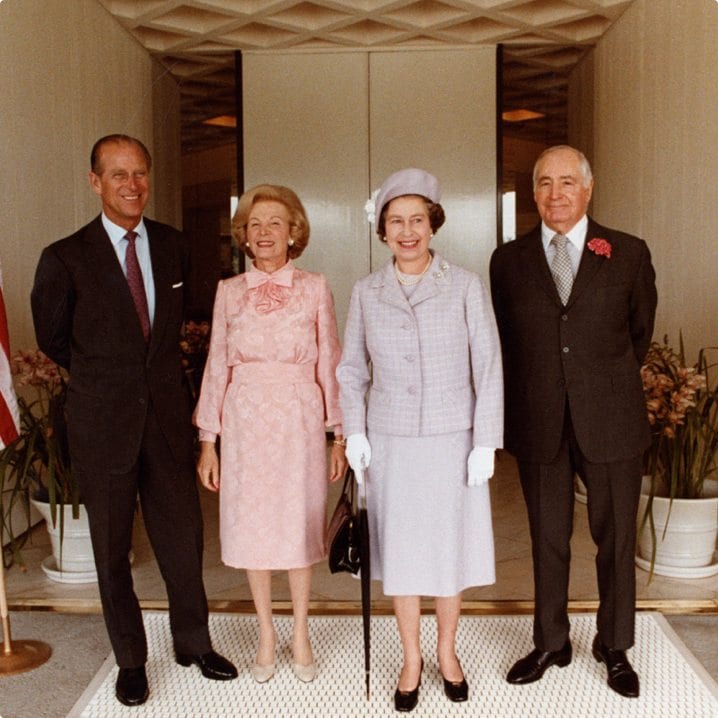 Four distinguished individuals posing for a photograph, two men wearing suits and two women in dresses and hats, indoors with a neutral backdrop.