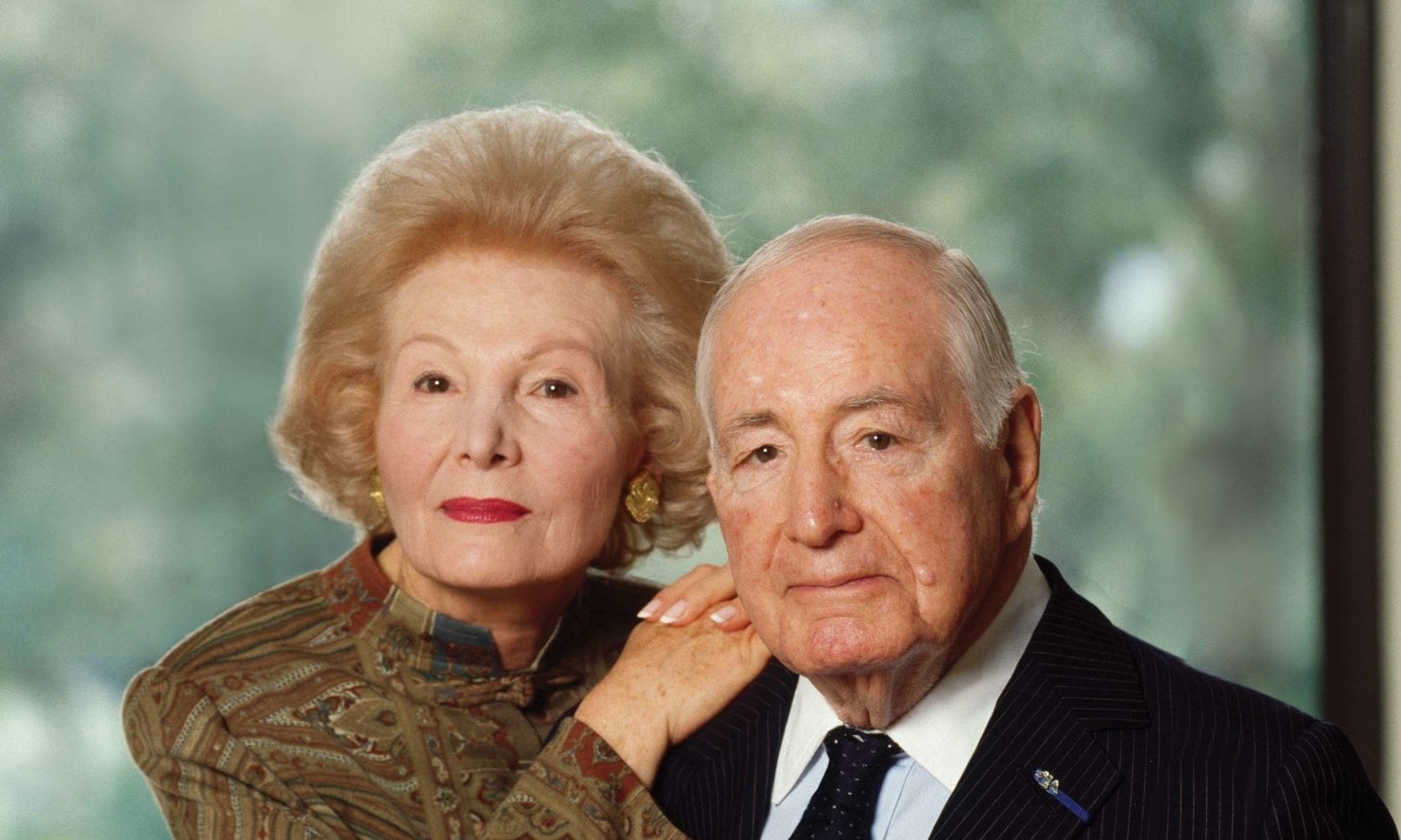 An elderly couple poses for a portrait with the woman standing slightly behind the man, resting her hand on his shoulder. Both are dressed formally, the woman in a paisley patterned dress with gold earrings and the man in a pinstripe suit with a tie and lapel pin. They appear dignified and composed against a blurred natural background.