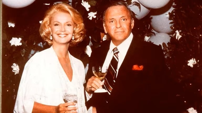 A smiling man and woman dressed in formal wear holding champagne glasses, with a background of balloons and flowers.
