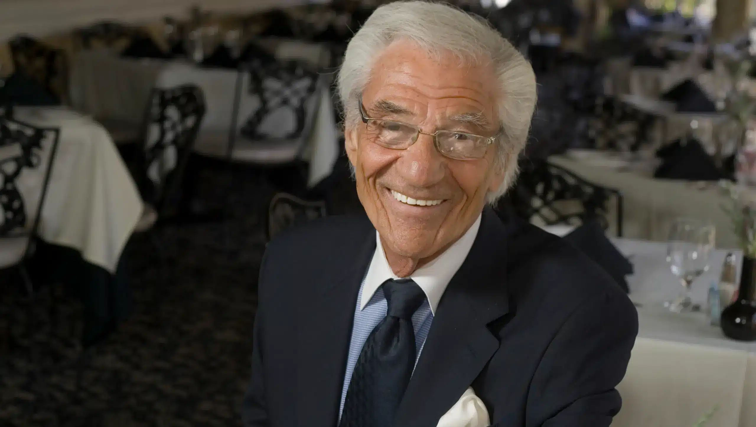An elderly man with white hair and glasses smiling at the camera, wearing a dark suit and tie, at an event with tables set in the background.