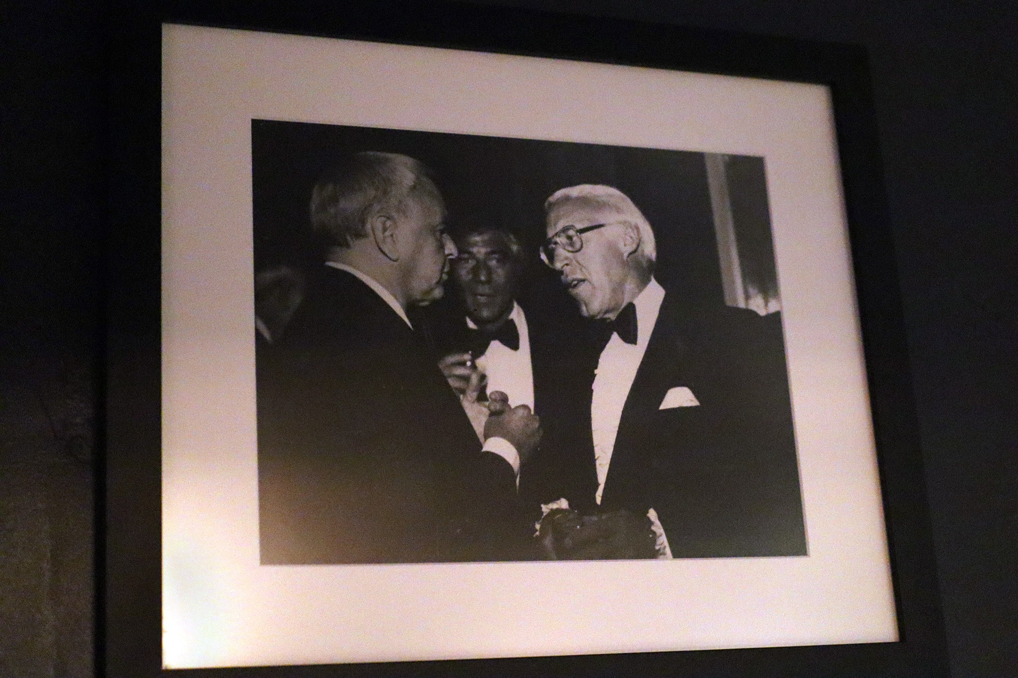 Black and white photograph displayed in a frame on a wall, capturing a moment where two older men in formal attire with bow ties are having a conversation. The man on the right appears to be gesturing with his hand while talking to the man on the left.