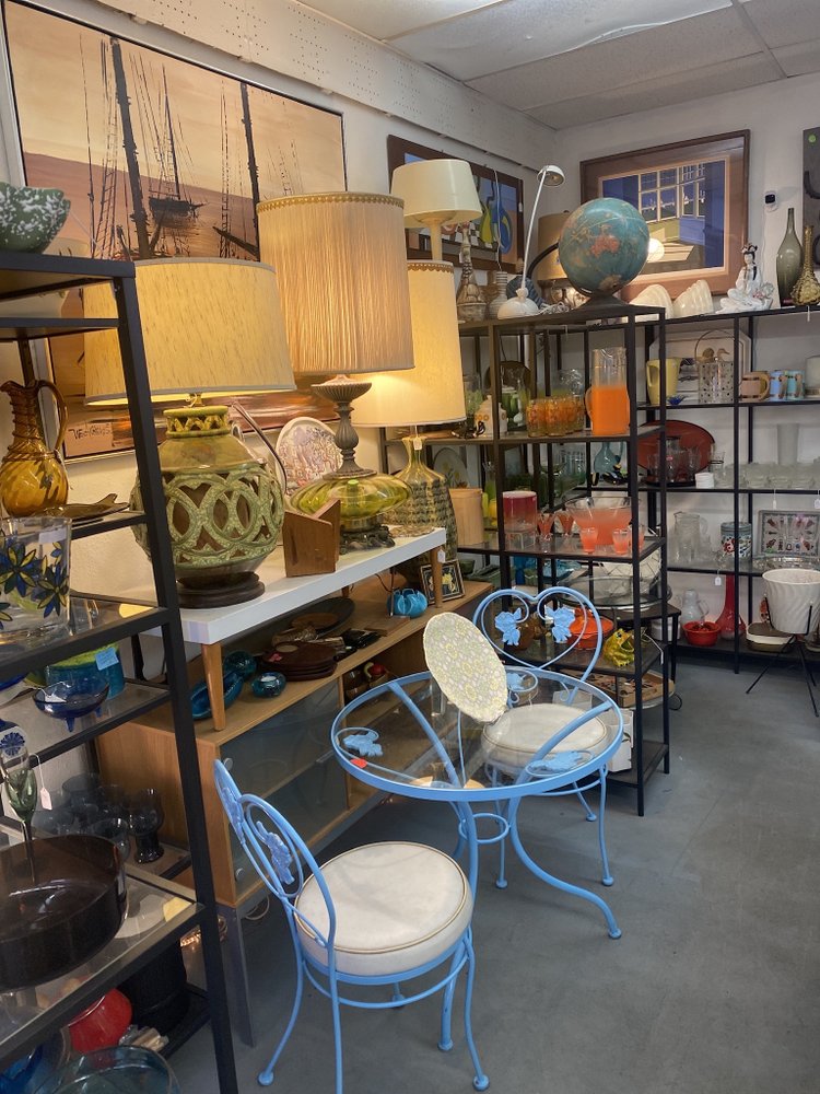 An antique shop interior with an assortment of items including lamps, a framed painting of ships, ceramic vessels, glassware, a globe, and a blue bistro table set with chairs.