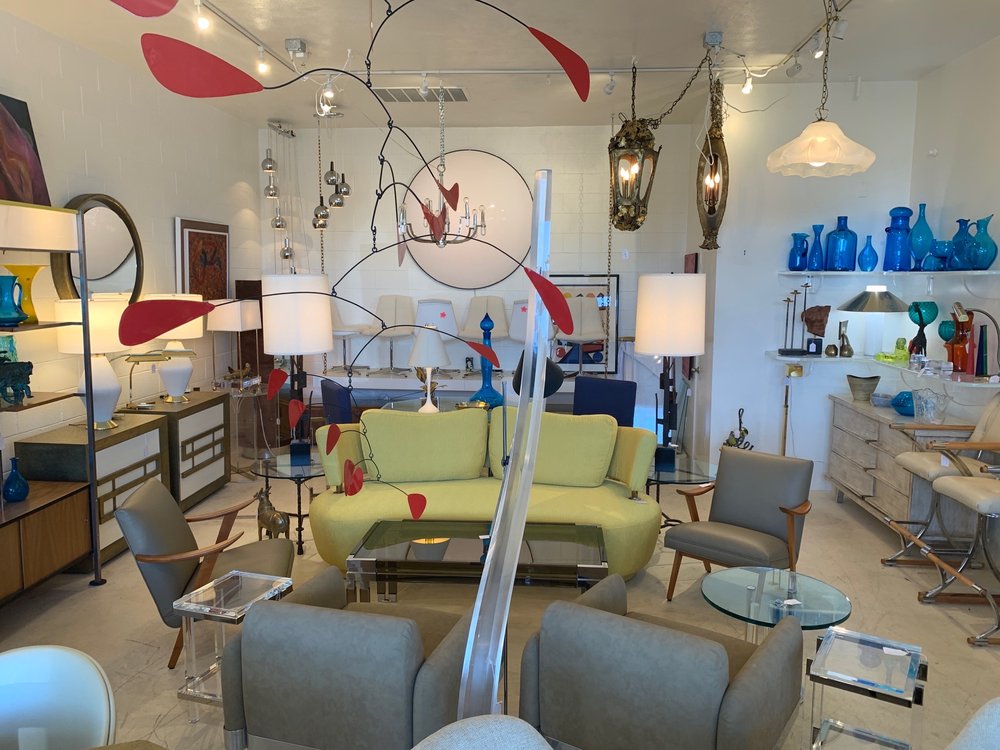 Alt text: A vintage furniture store interior with various mid-century modern pieces, including sofas, chairs, tables, and colorful glassware displayed on shelves. A distinctive mobile with red elements hangs from the ceiling amidst diverse lighting fixtures.