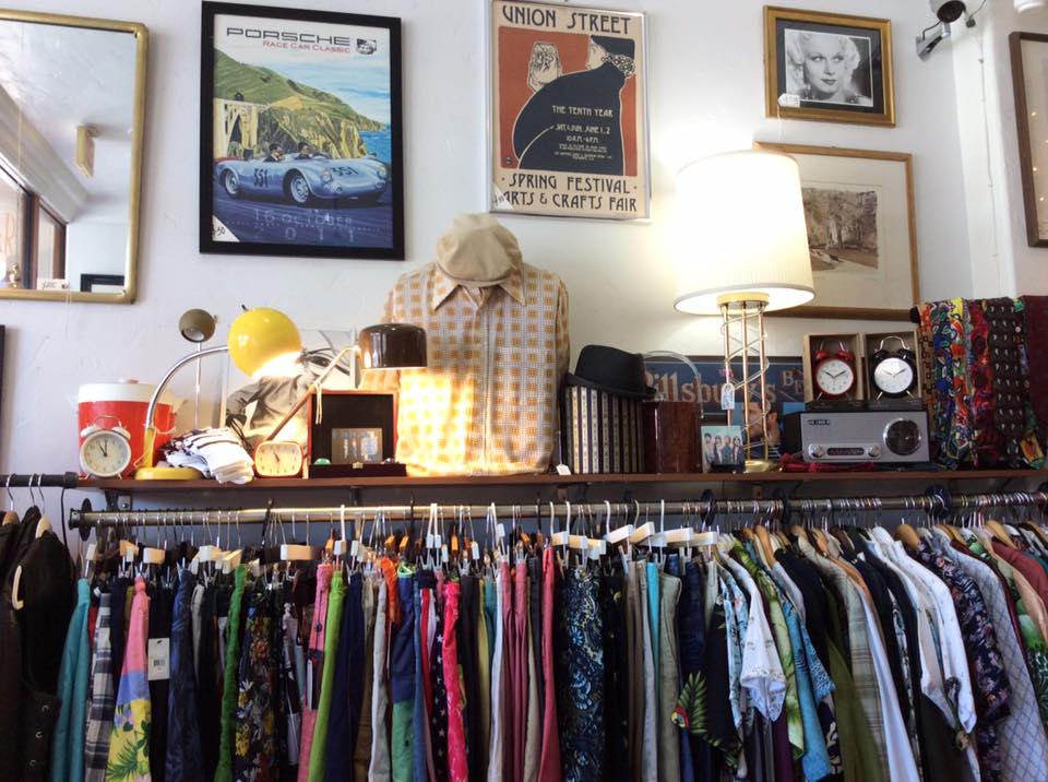 A vintage boutique interior with a variety of clothes hanging on racks, assorted retro objects on shelves, and vintage posters on the walls.