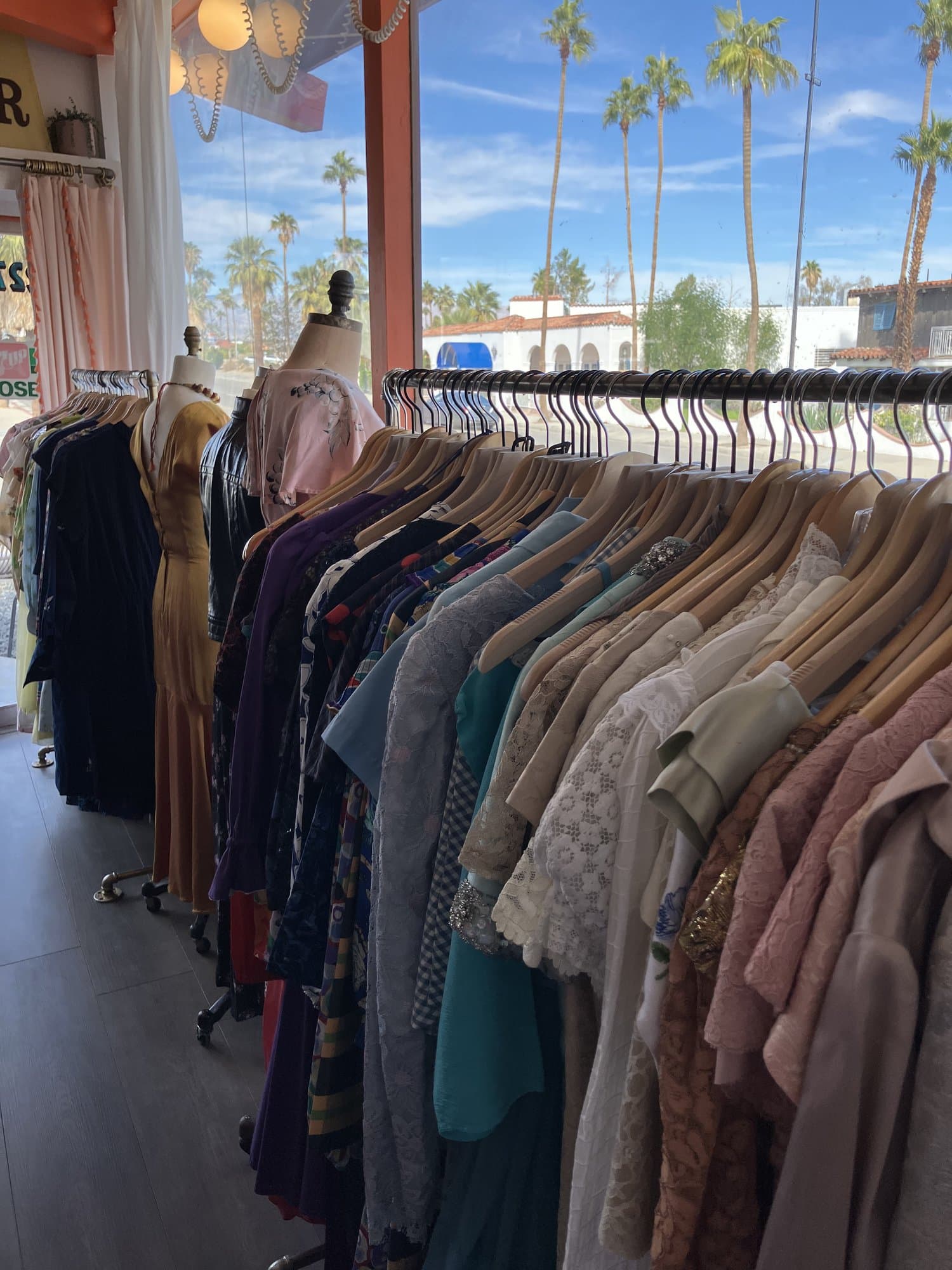 An interior view of a boutique with racks of various colorful dresses and garments, two mannequin torsos displaying clothes at the end of the rack, and a sunny view of palm trees through the front window.