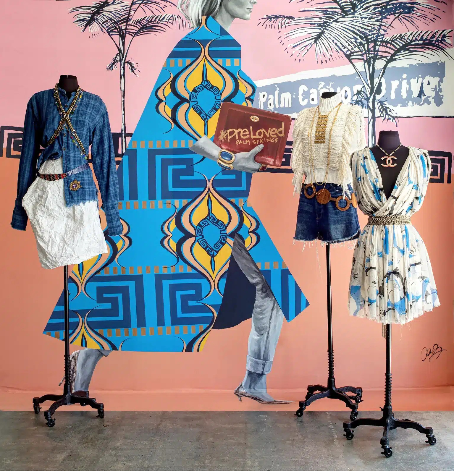 A display of three mannequins with stylish outfits against a colorful mural wall, featuring geometric patterns and a partial illustration of a woman's silhouette. The center mannequin is holding a sign that says "#preLoved PALM SPRINGS."