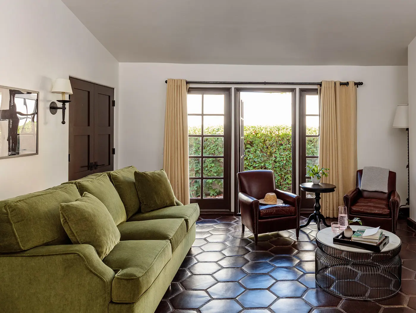 A cozy living room with a green velvet sofa, two brown leather armchairs, hexagonal tile flooring, and French doors opening to greenery. Decor includes a wall sconce, window curtains, a round coffee table with books, and a side table with a vase.