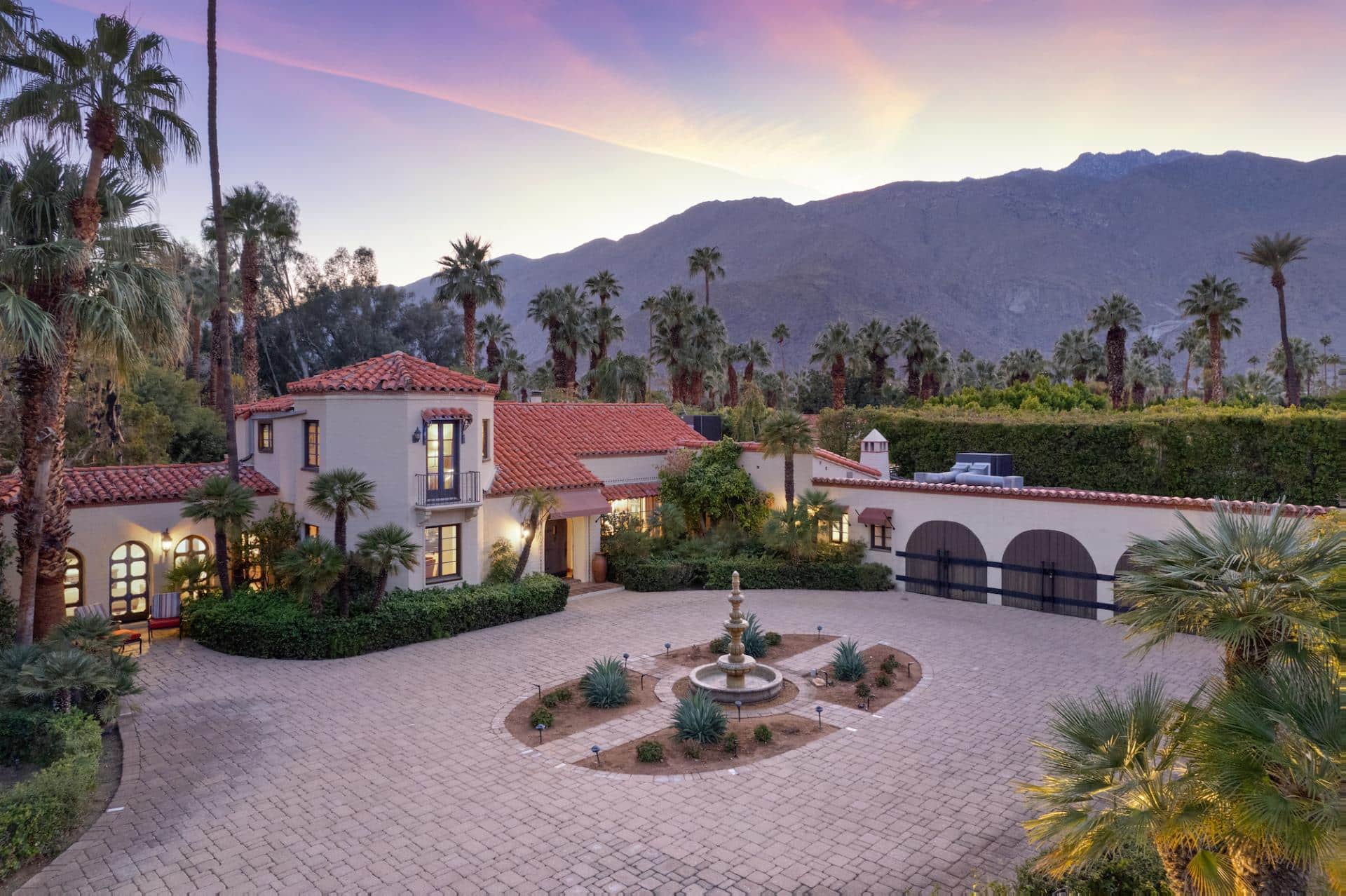 Luxurious Spanish-style villa with red-tile roofs, a fountain in a circular driveway, surrounded by palm trees, with a backdrop of mountainous terrain under a sunset sky.