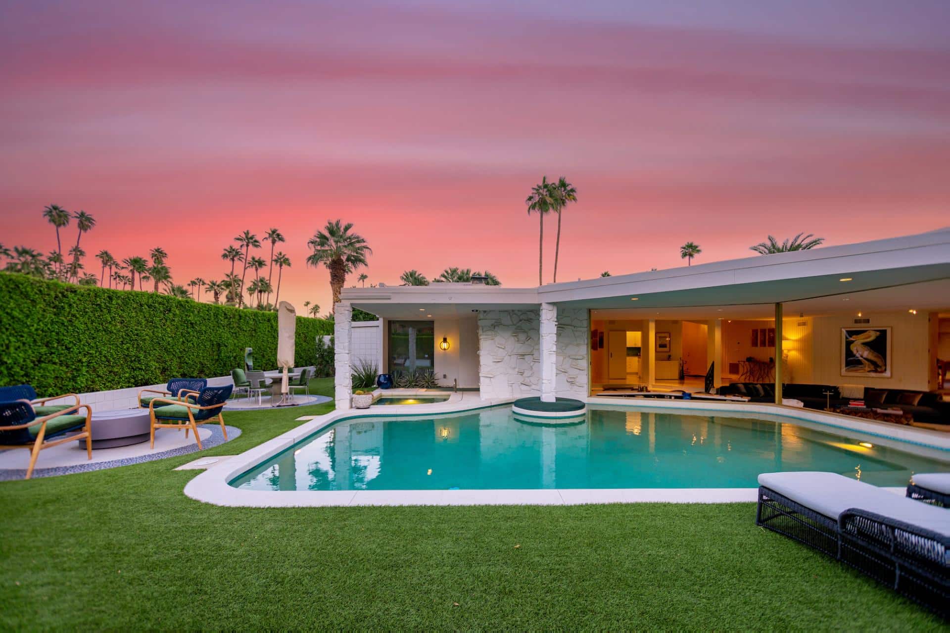 A luxurious modern house with an outdoor swimming pool at twilight, surrounded by palm trees against a pink and orange sky.