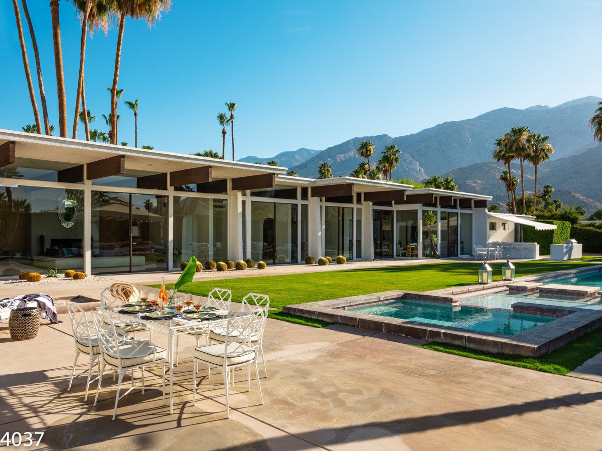 A modern luxury home with large glass windows, an outdoor dining area set up for a meal, a swimming pool in the foreground, surrounded by well-manicured lawns and tall palm trees, with mountains in the background under a clear blue sky.