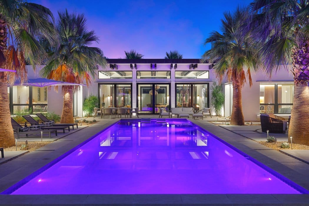 A modern house at twilight with an illuminated blue swimming pool in the foreground, surrounded by palm trees and outdoor furniture.