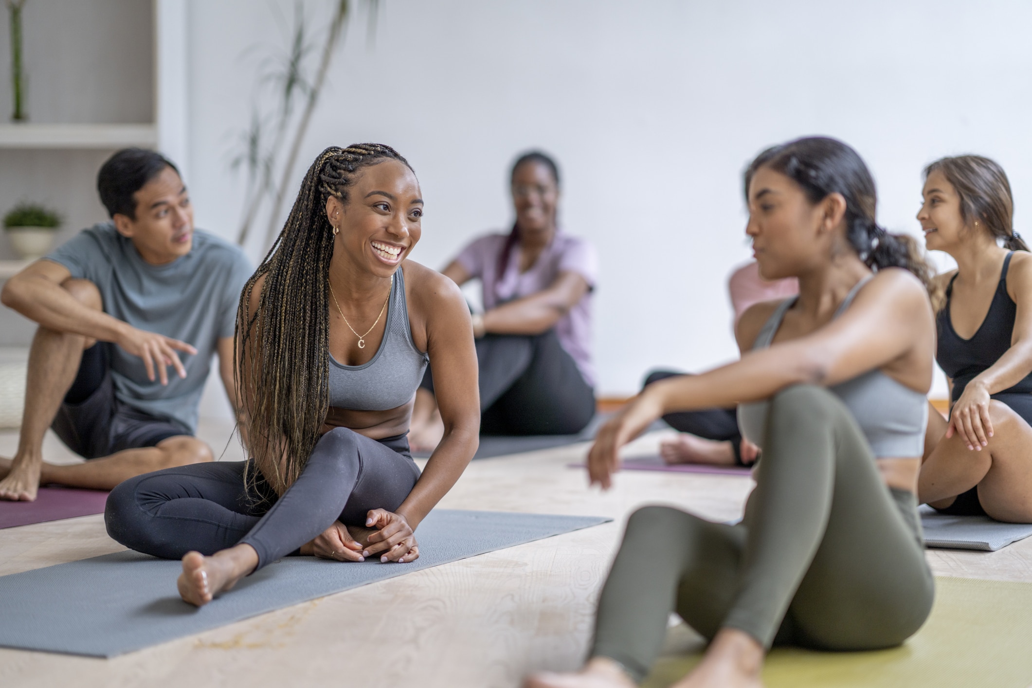 A small group of mature adults sit on individual yoga mats as they take a break after their fitness class. They are each dressed casually in athletic wear and are smiling as they talk amongst themselves.