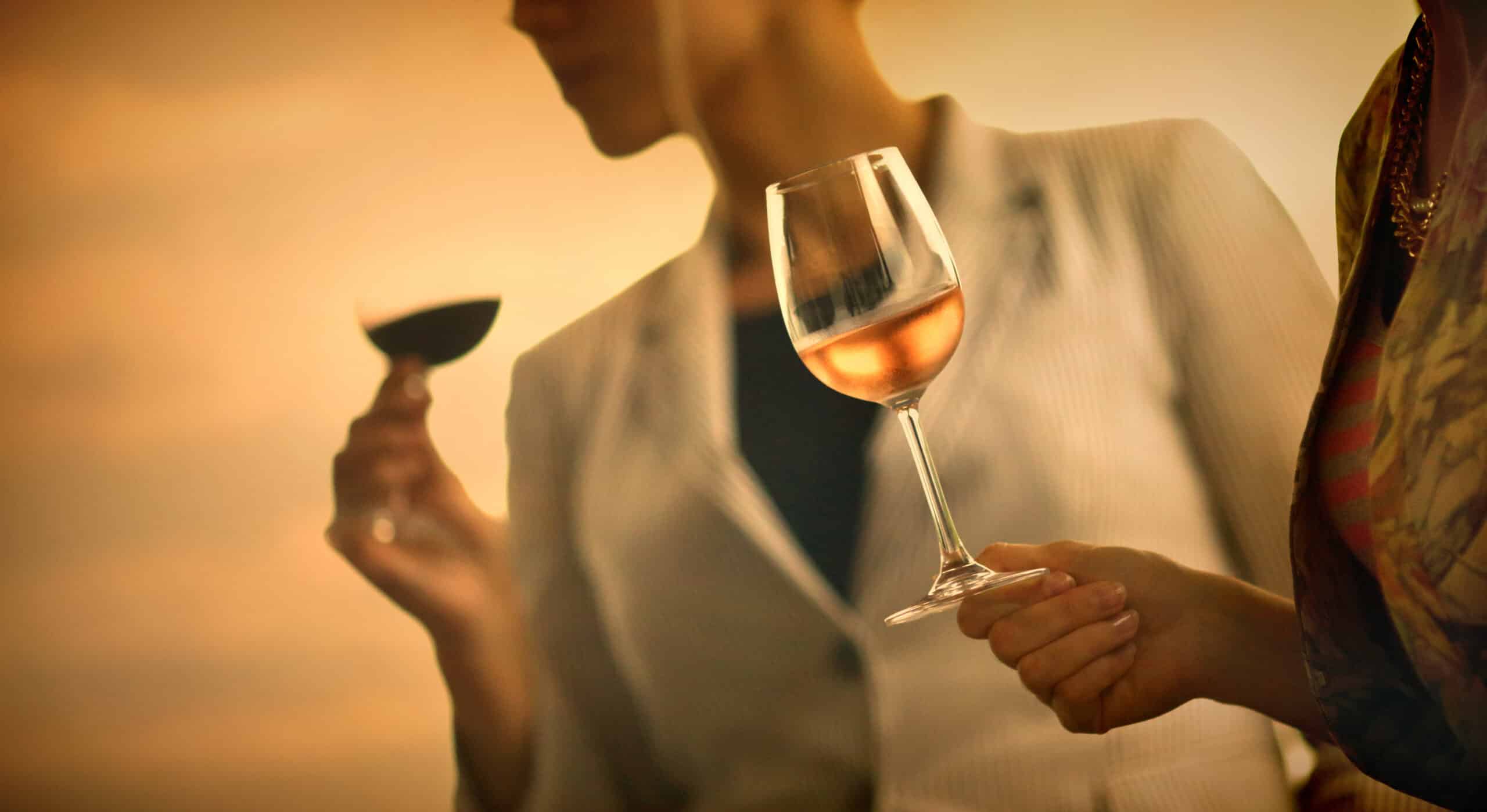 Group of unrecognizable women tasting wine . Comparing appearance, smell, aroma,t aste, aftertaste, holding wineglasses on the stems.The closest person to he camera is in focus is holding a glass of red wine and looking at its color. It's summer sunset, image is orange toned.