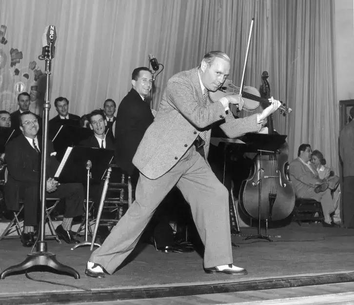 American actor, entertainer and comedian Jack Benny plays the violin on stage with an orchestra and a conductor during a radio broadcast from the Plaza Theatre in Palm Springs, circa 1941. 