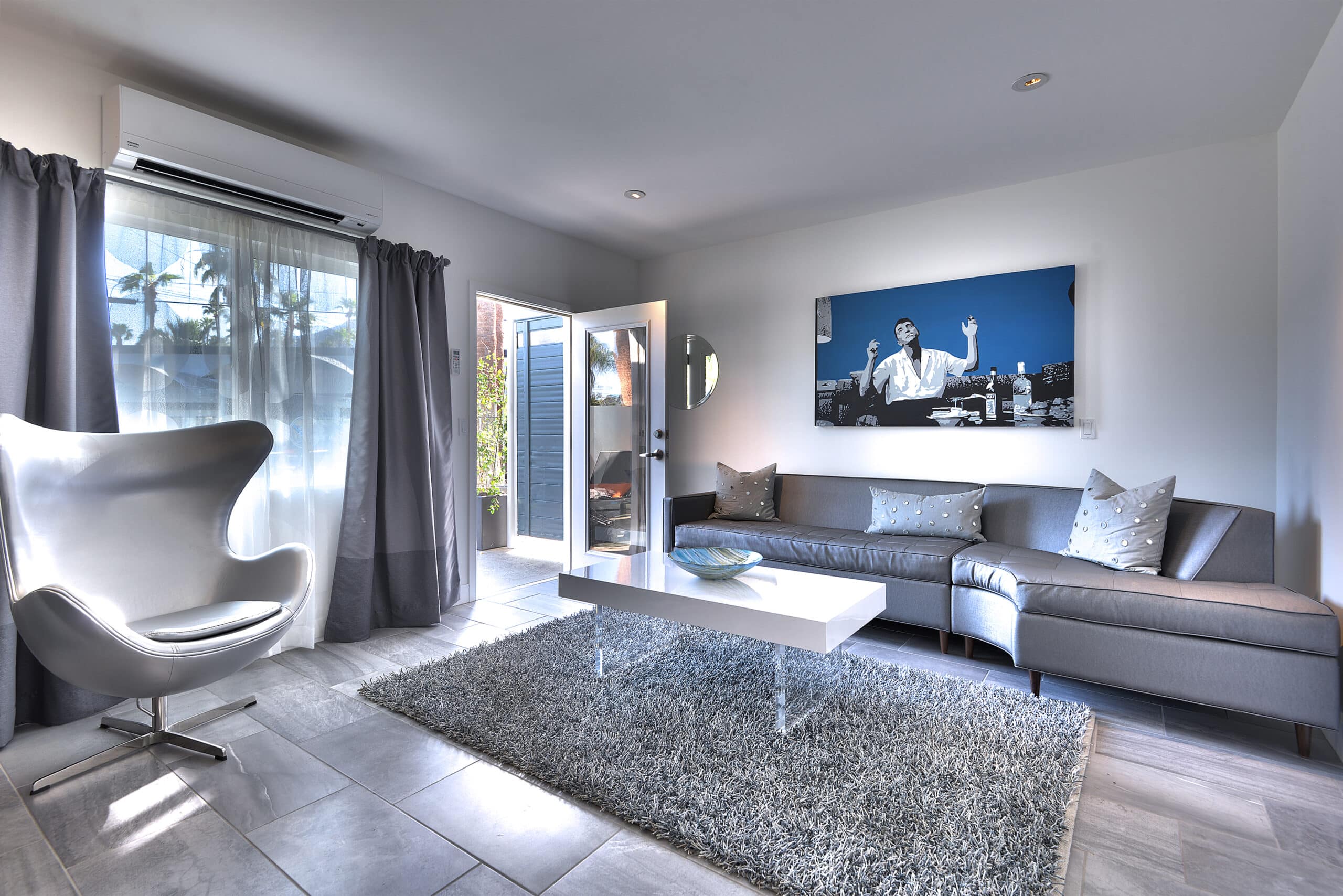 A modern living room with a sectional gray sofa, white designer chair, shaggy area rug, and a large monochromatic wall art above the couch. Glass doors lead to the exterior, and the room is brightly lit with natural light.