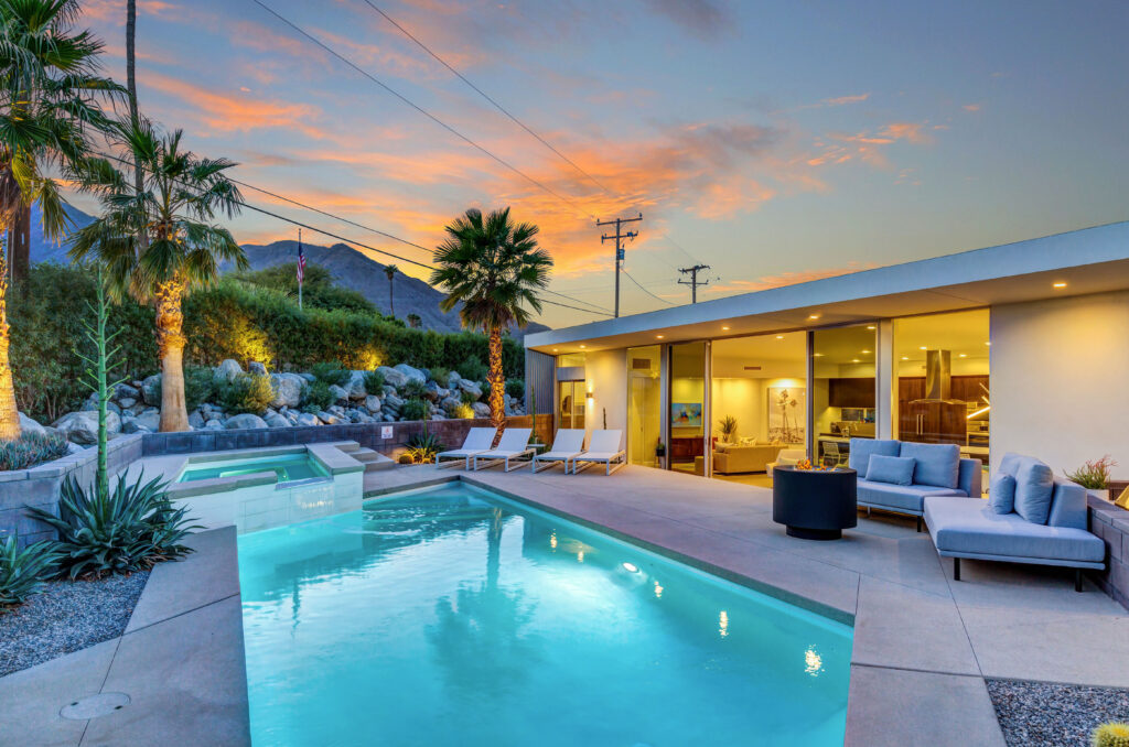 Modern backyard with a swimming pool at dusk, featuring outdoor furniture, palm trees, and a mountain view with a colorful sky.