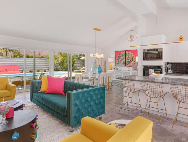 A bright and colorful modern living room with a high ceiling, a teal sofa with pink pillows, two yellow armchairs, and a black coffee table. In the background, there is an open kitchen with bar stools and a dining area, with a view of a pool outside through large windows. The room is accented with vibrant artwork and eclectic decor.