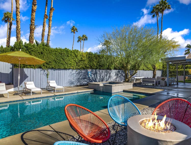 An outdoor pool area with lounge chairs and a yellow umbrella, surrounded by palm trees and greenery, featuring colorful chairs and a fire pit in the foreground.