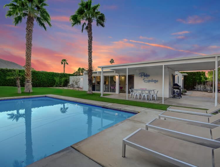 A backyard with a swimming pool at twilight, framed by palm trees and featuring a house with the text "Palm Springs" on the exterior wall, lounge chairs, and a dining area under a shelter.