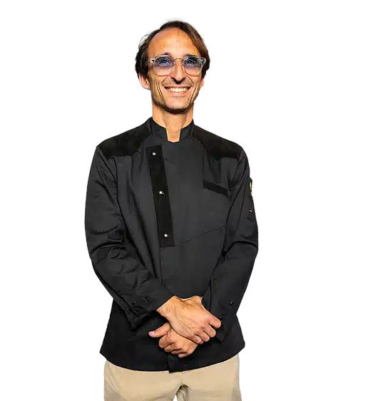 A man with a cheerful expression wearing a black shirt and beige pants, with his hands clasped in front of him. He has slim-framed glasses and short brown hair. The image background is transparent with some graphical elements.