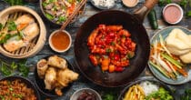 A variety of Asian dishes spread out on a table, including spring rolls in a bamboo steamer, a bowl of salad with beef, white rice with sesame, spicy red chicken with vegetables in a black skillet, and other small dishes containing ingredients like lettuce, herbs, and sauces.