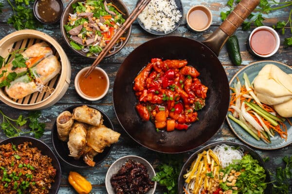 A variety of Asian dishes spread out on a table, including spring rolls in a bamboo steamer, a bowl of salad with beef, white rice with sesame, spicy red chicken with vegetables in a black skillet, and other small dishes containing ingredients like lettuce, herbs, and sauces.