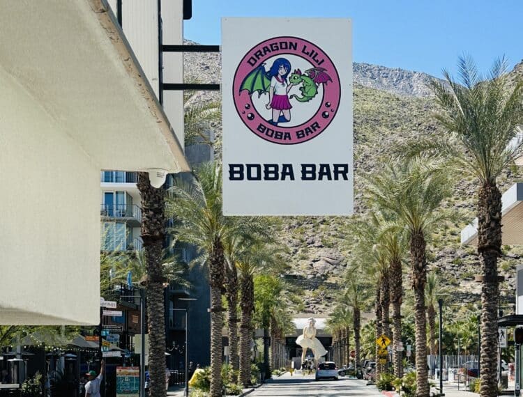 A sunny pedestrian street lined with palm trees, featuring a sign for "Dragon Lily Boba Bar" on the left, with a mountain background and a sculpture visible in the distance.