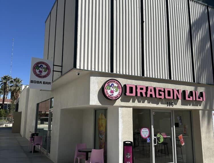 A boba tea shop named "DRAGON LILI" with pink signage and outdoor seating area featuring two pink chairs and a table, located on a sunny street with palm trees in the background.