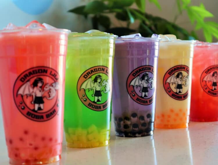 Five colorful boba tea drinks in clear plastic cups with a logo featuring a dragon and a person, displayed in a row with assorted toppings visible at the bottom of each cup.