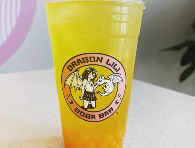 A clear plastic cup of yellow bubble tea with orange tapioca pearls, ice cubes, and the logo "DRAGON LILY BOBA BAR" featuring an illustration of a girl with dragon wings and a small dragon.