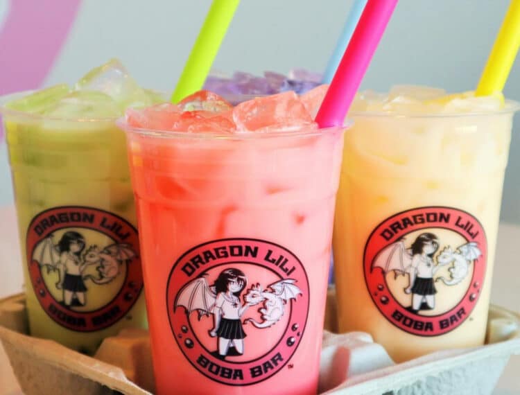 Three colorful bubble tea drinks in clear plastic cups, with the logo "DRAGON LILY BOBA BAR" printed on them, are placed in a cardboard drink carrier. Each drink has a different colored straw - green, pink, and yellow - and seems to contain ice with varying shades of green, pink, and yellow liquid.