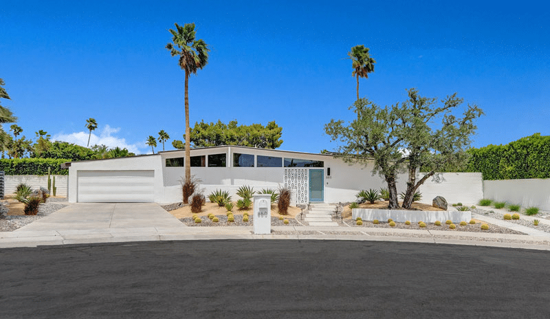A single-story white mid-century modern house with a flat roof, large windows, a two-car garage, and a landscaped front yard with palm trees, under a clear blue sky.