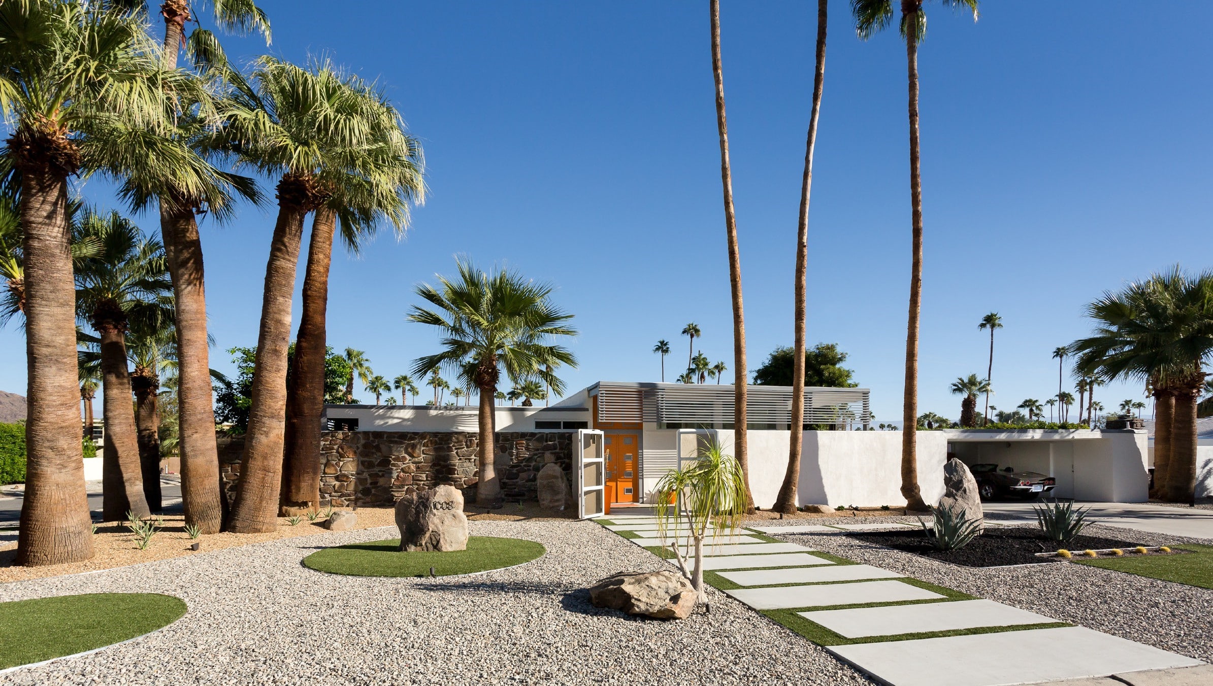 Modern house with a flat roof and large windows, flanked by tall palm trees, featuring a landscaped front yard with gravel, green grass accents, and stepping stones leading to an orange front door.