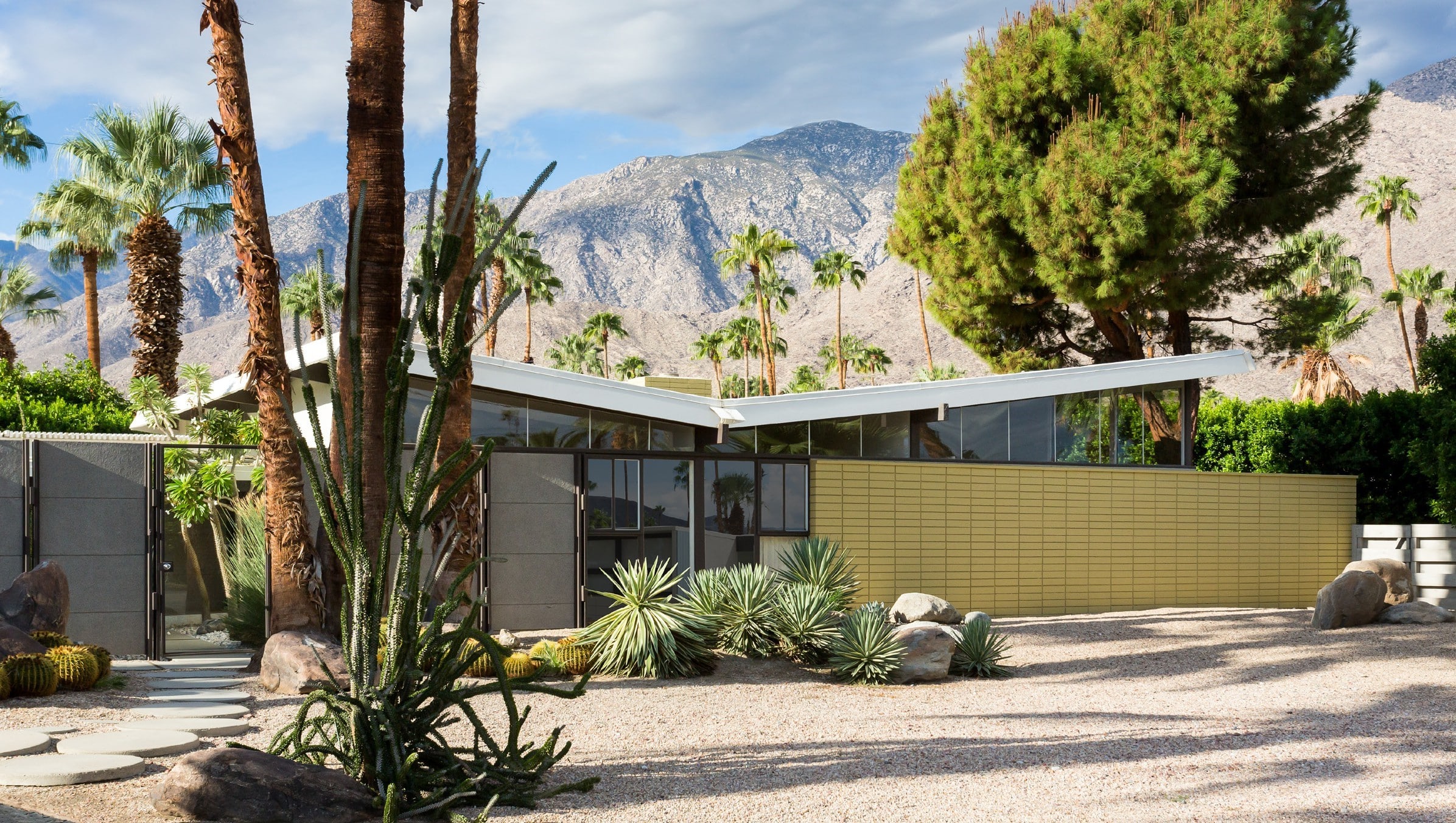 A mid-century modern house with a flat roof and large windows, surrounded by desert landscaping including cacti, palm trees, and rocks, set against a backdrop of mountainous terrain.