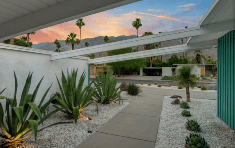 Modern house exterior with desert landscaping including agave plants, viewed at dusk with pastel-colored sky and mountains in the background.