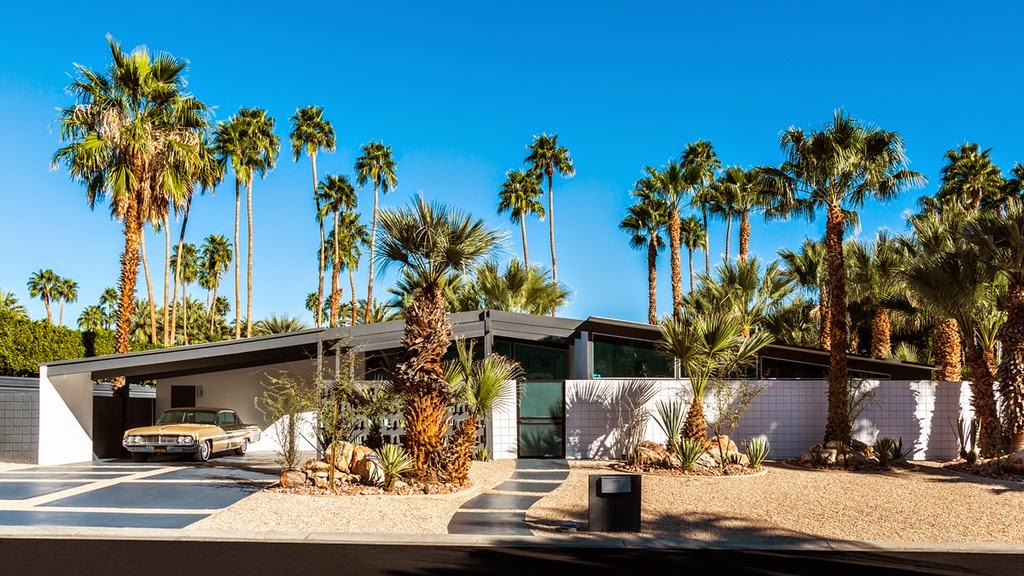 A mid-century modern house with a flat roof and large windows, surrounded by tall palm trees. A vintage car is parked under the carport. The home features a mix of clean lines and geometric shapes, with a desert landscaped front yard.