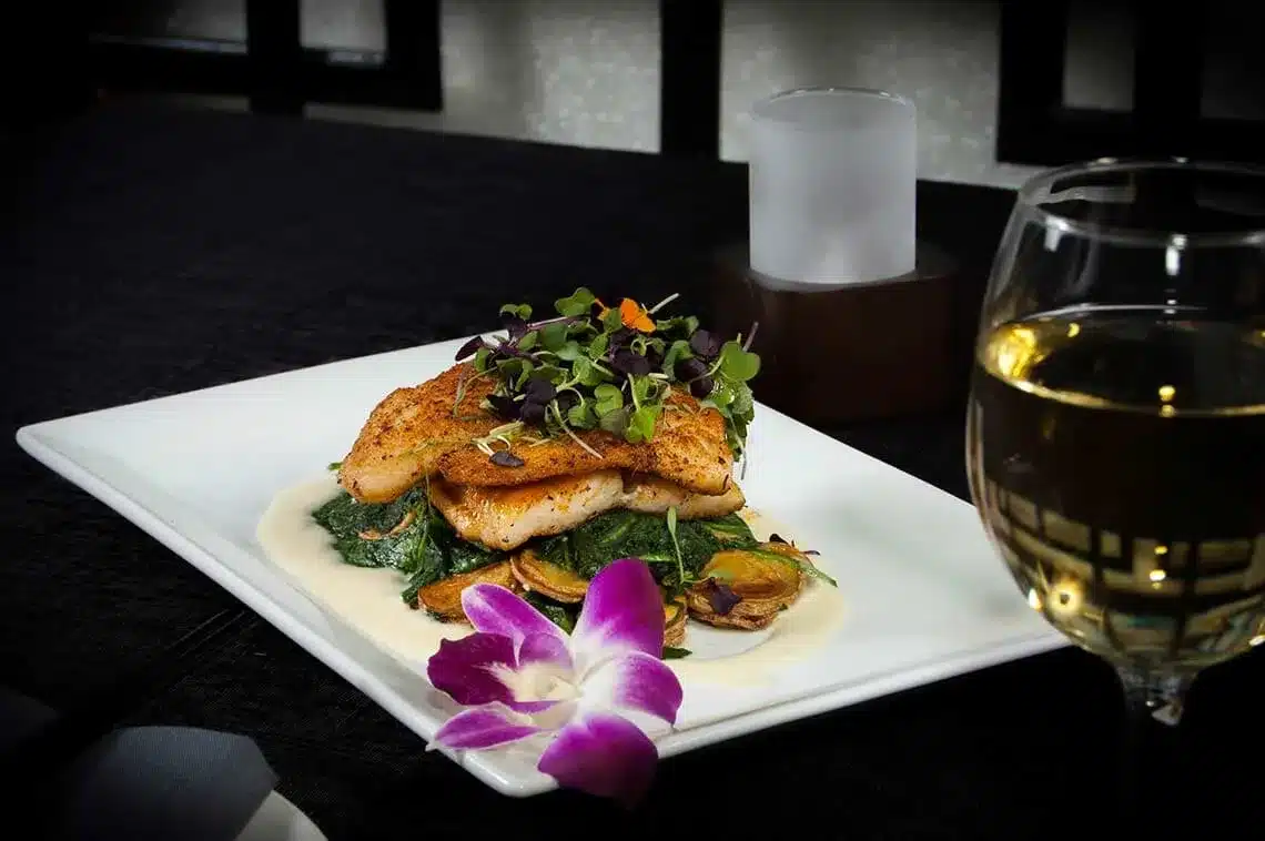 Seared fish fillet on a bed of spinach with microgreens garnish, served on a white plate with a decorative flower, accompanied by a glass of white wine.