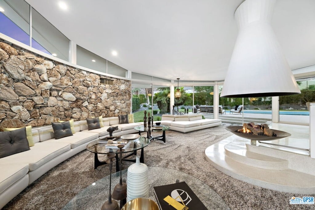 A modern living room with large stone wall feature, white sectional sofas, a unique central fireplace, glass walls with a view of a pool, and polished decor.