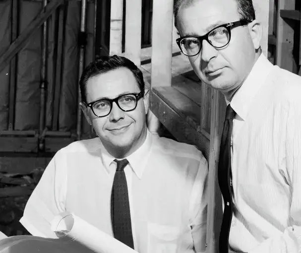 Two men wearing glasses and dress shirts with ties, one holding architectural plans, standing together in a room with wooden framing in the background. The image is in black and white.