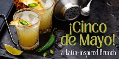 Two glasses of a citrus cocktail garnished with chili slices and a pineapple piece on a tray with a lime wedge, whole jalapeño, cocktail shaker, and ice cubes, with the text "¡Cinco de Mayo! a latin-inspired Brunch" on the right side.
