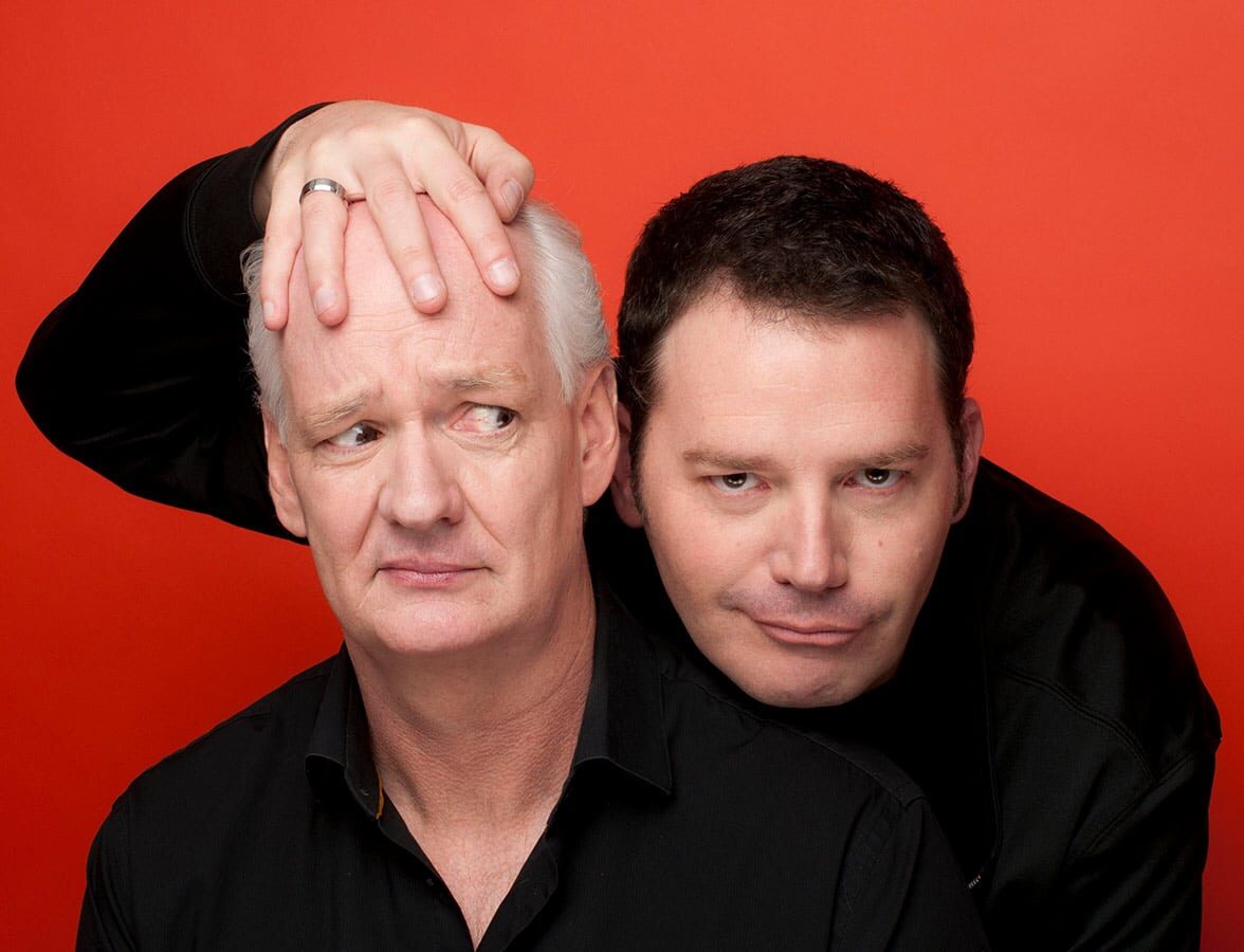 Two men posing closely with one man resting his chin on the other's head against a red background.