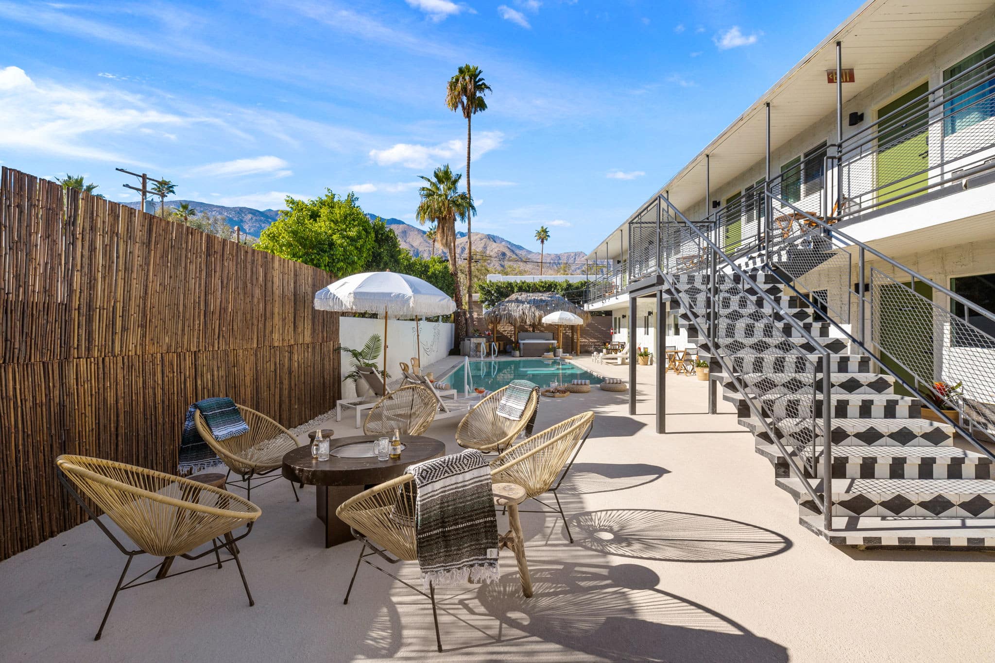 A sunny outdoor patio with wicker chairs and tables, a white privacy fence, and an adjacent two-story building with an exterior staircase. Behind the area is a pool with loungers and umbrellas, set against a backdrop of palm trees and mountains under a blue sky.