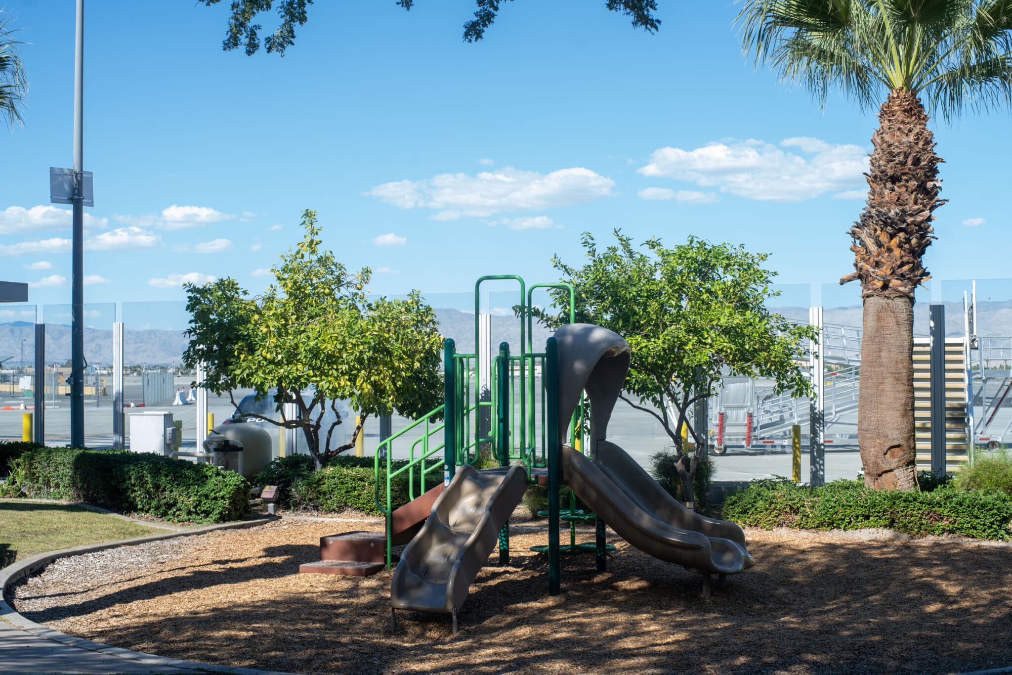 A playground with two slides and green railings, surrounded by trees and wood chips, with a palm tree on the right and mountains in the distance under a blue sky.