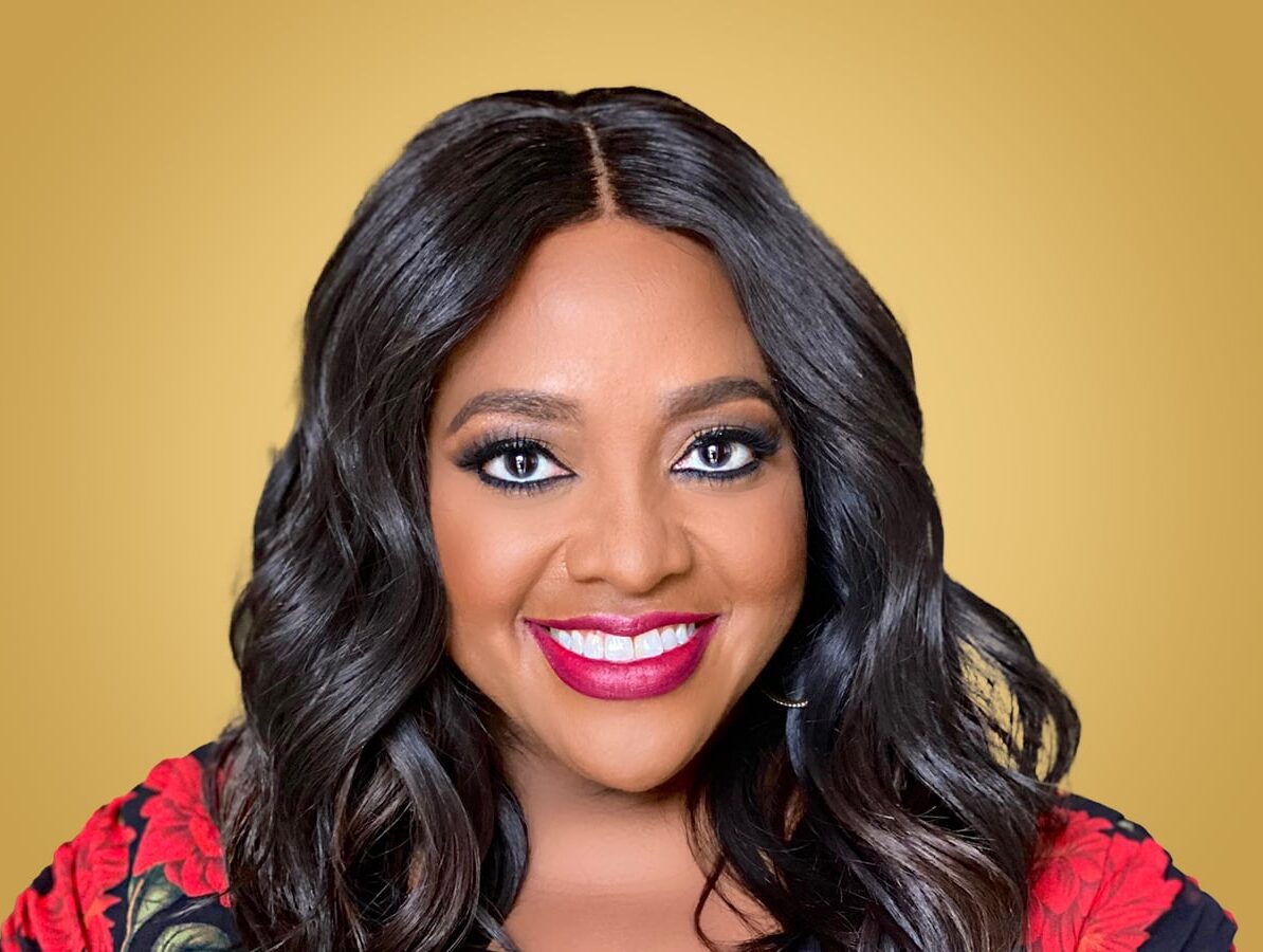 Portrait of a smiling woman with long wavy hair, wearing makeup with dark eyeshadow and red lipstick, and a floral top, against a yellow background.