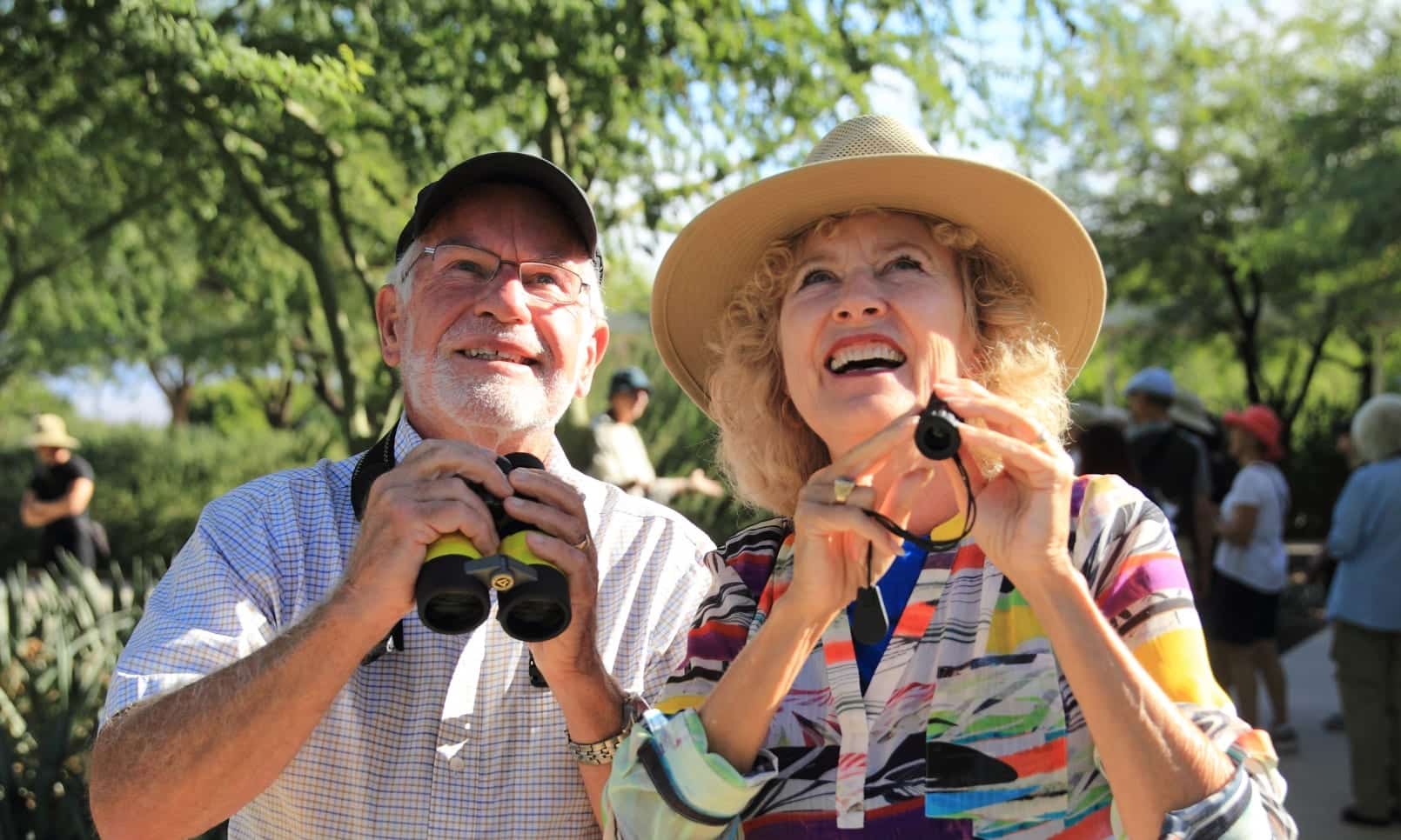 An older couple outdoors smiling with binoculars, the man wearing a cap and the woman in a sunhat, with other people and greenery in the background.