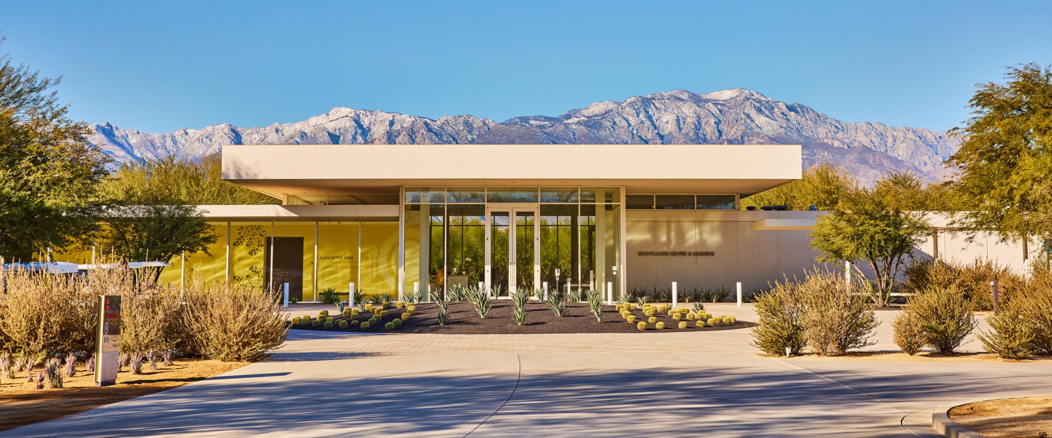 A modern building with large glass windows and a flat roof, with the text "A PLACE AT THE TABLE" on the front, set against a backdrop of towering mountains and a blue sky. The landscape features desert vegetation and a clear driveway leading to the entrance.