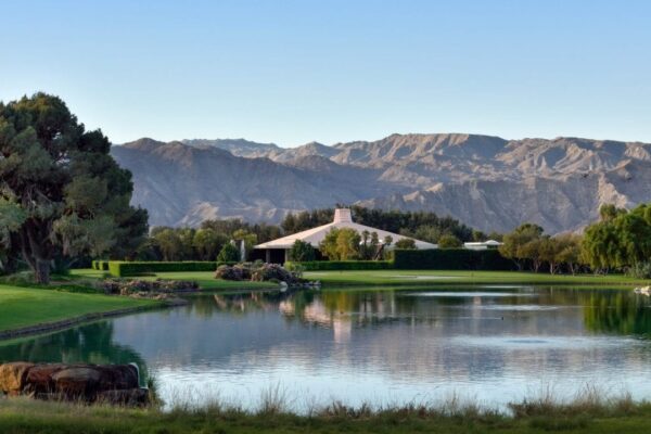 A tranquil golf course with a small pond reflecting the surrounding trees and a clear view of distant mountains against a blue sky.